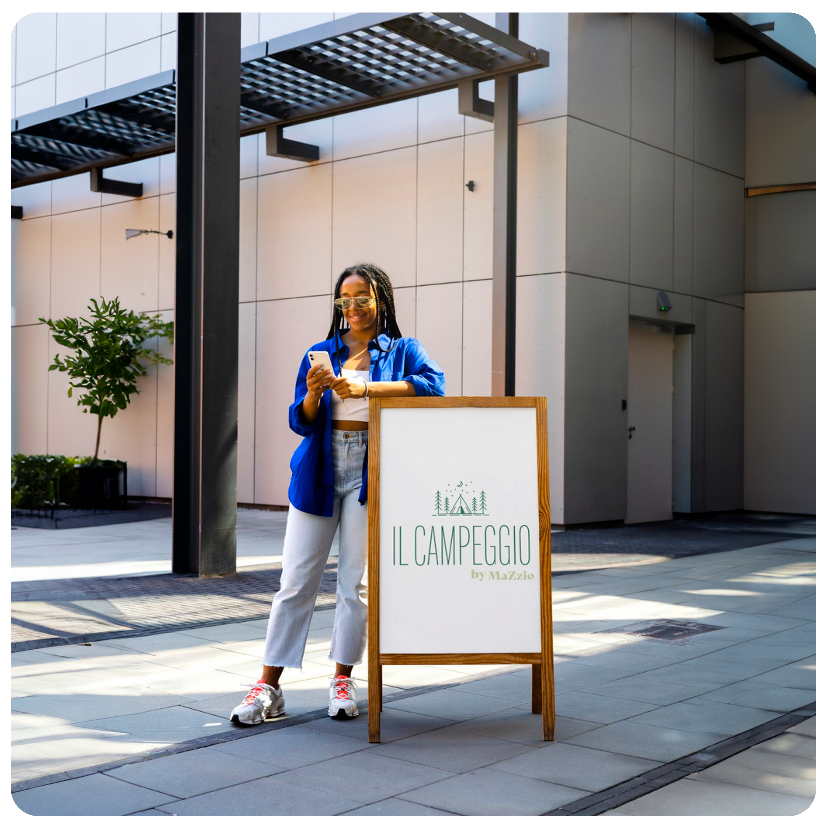 Une femme avec des lunettes de soleil et une chemise bleue est accoudée sur un stop trottoir en bois de couleur marron clair, dans une rue regardant son téléphone